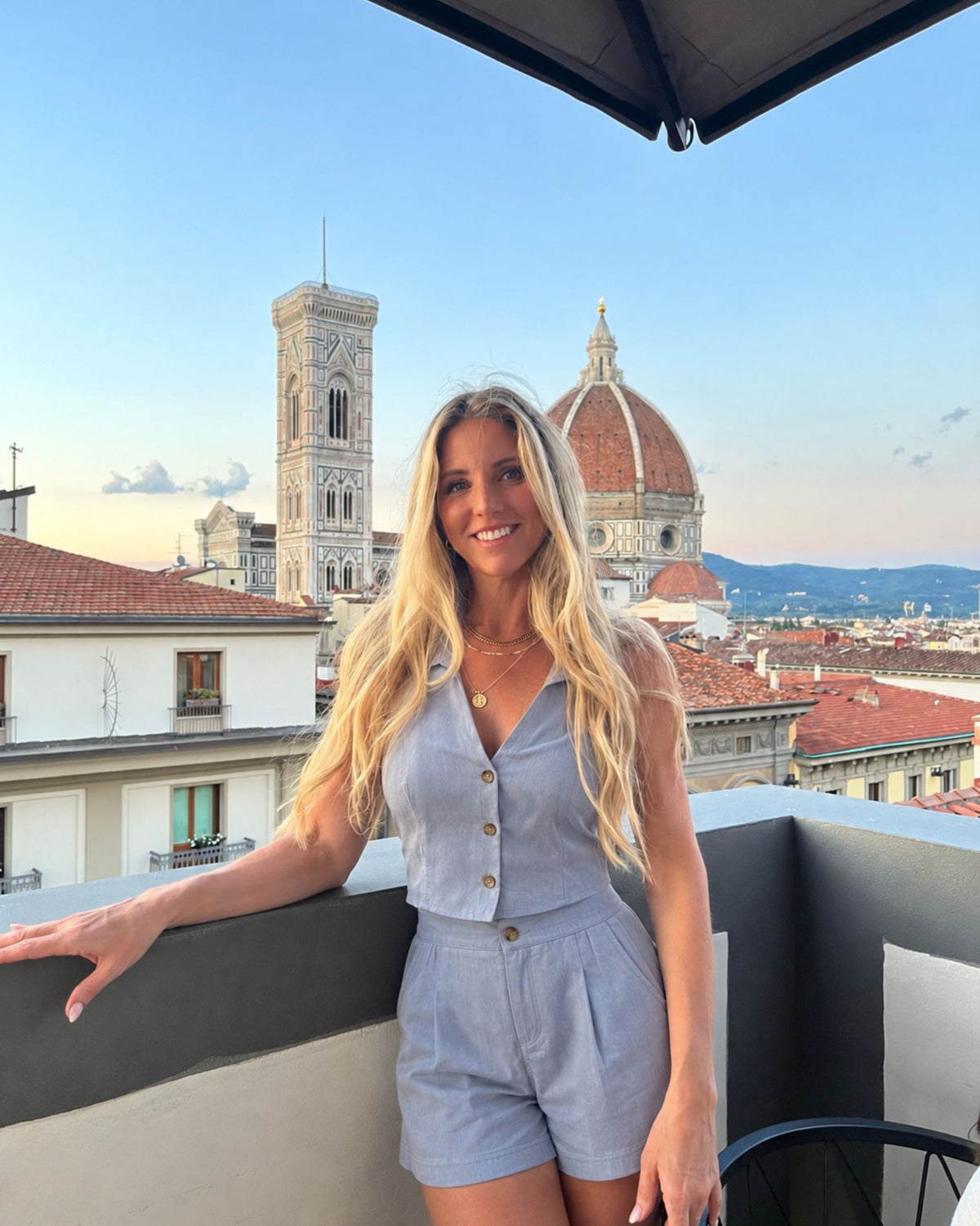 A smiling blonde woman stands on a rooftop balcony wearing a light blue sleeveless button-front romper with matching shorts, with Florence's skyline and the Florence Cathedral dome in the background.